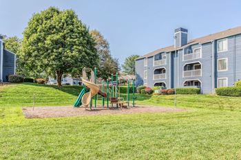 A playground with a slide and a green tree in front of a building.